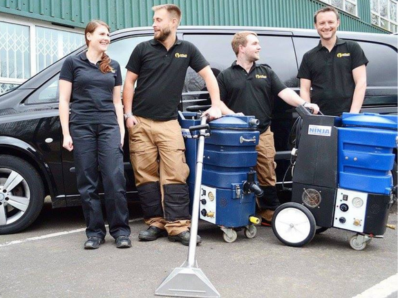 This is a photo of Paul's Cray Carpet Cleaning carpet cleaners (three men and one woman) standing in fromt of their black van, with two steam cleaning carpet machines next to them works